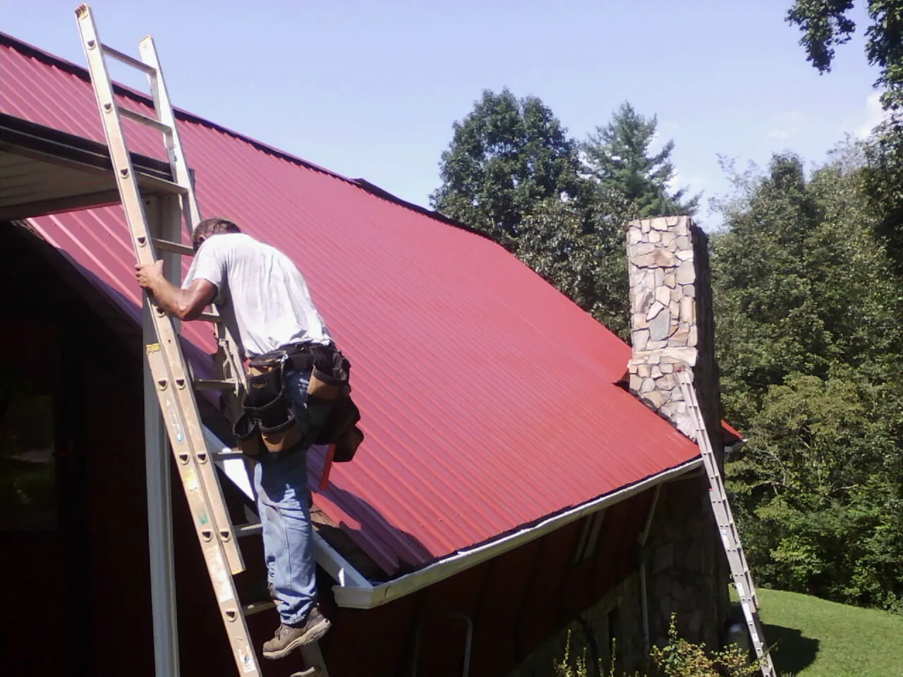 Roofer on a ladder beside a red metal roof and stone chimney — Soffit & Fascia Repair by Roofing Brothers in Dowelltown